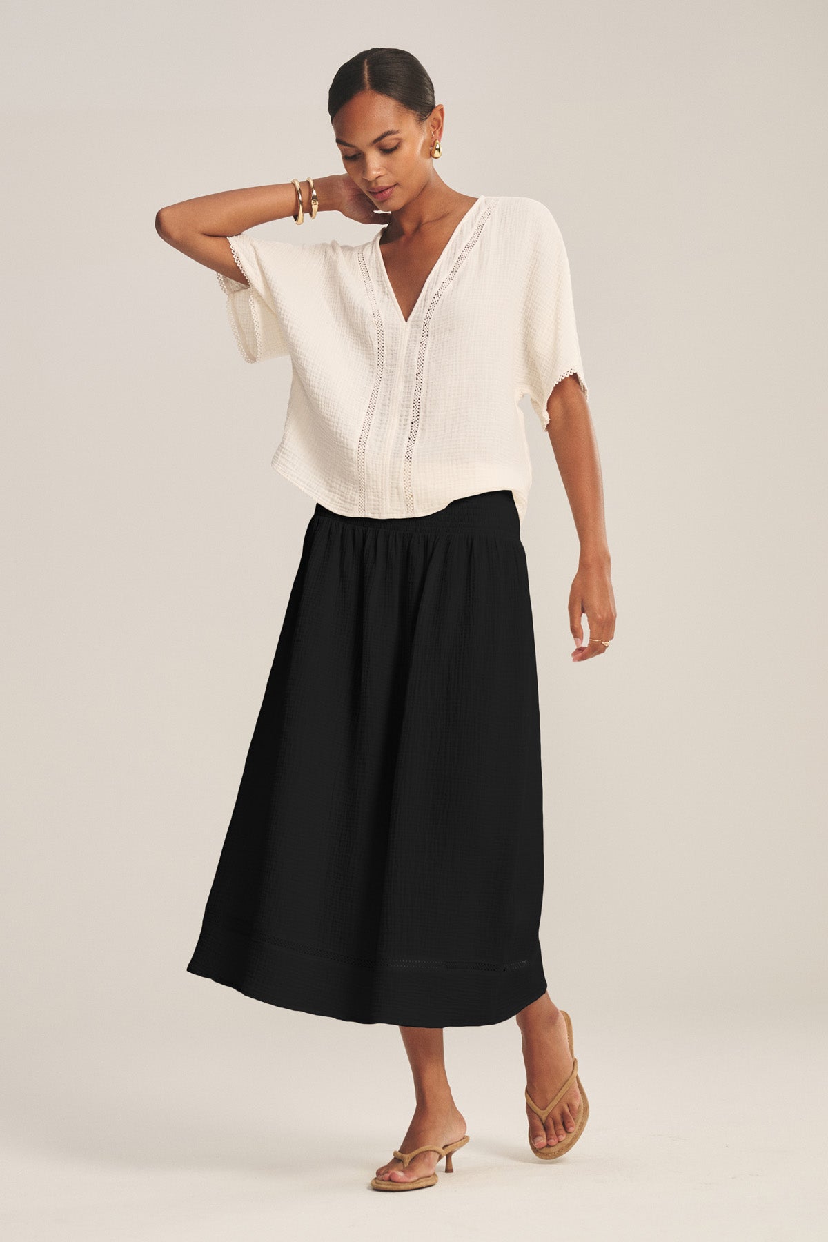 A woman stands against a plain background wearing a loose white short-sleeve blouse, tan sandals, and the MAZIE SKIRT by Velvet by Graham & Spencer—a black skirt with a smocked waist.-41296920084673