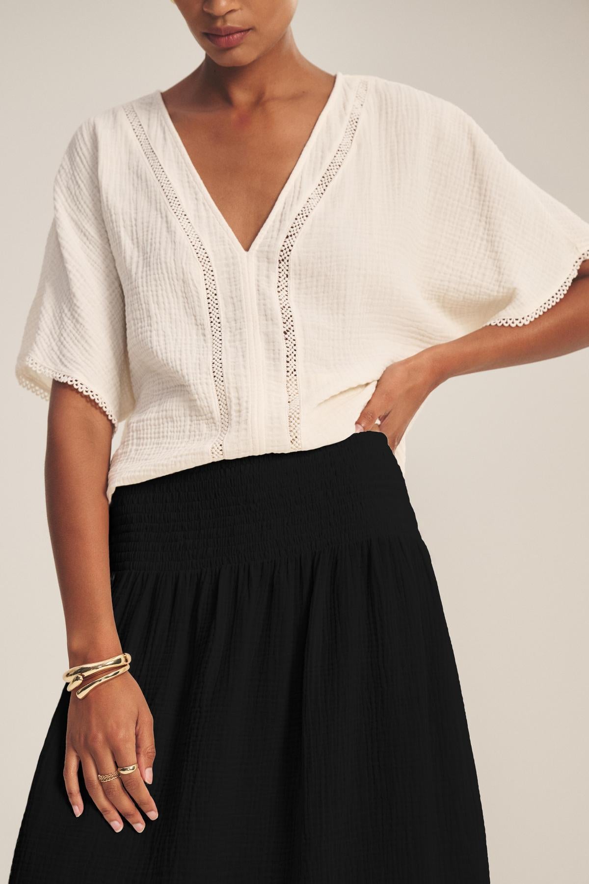 A person wears a white textured short-sleeve blouse with lace trim and the MAZIE SKIRT by Velvet by Graham & Spencer in black cotton gauze, accessorized with gold bracelets and rings.