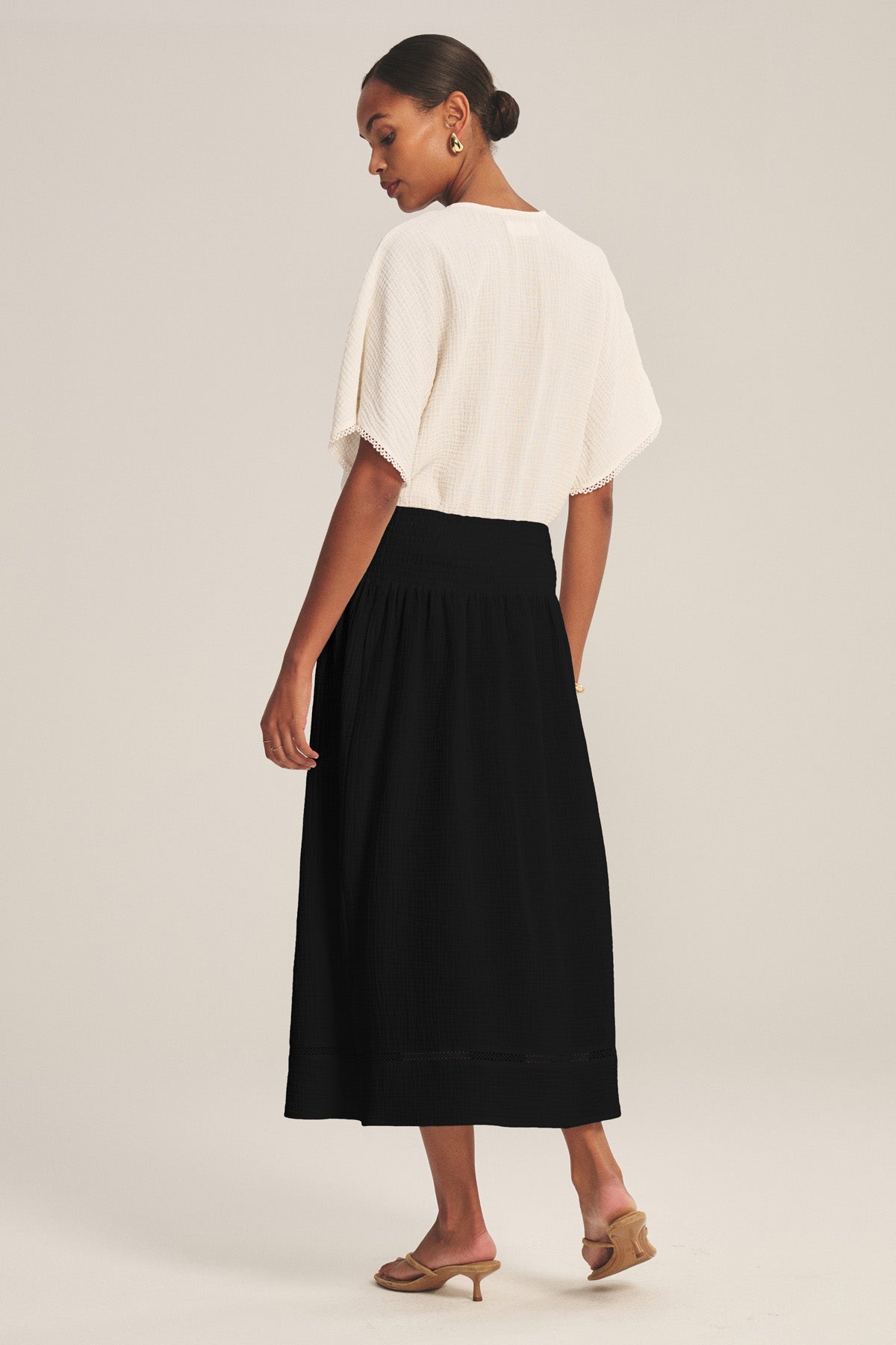 A woman in profile faces left, wearing a white textured short-sleeve top, the MAZIE SKIRT by Velvet by Graham & Spencer with a black lace hem, tan heeled sandals, and hoop earrings against a plain background.