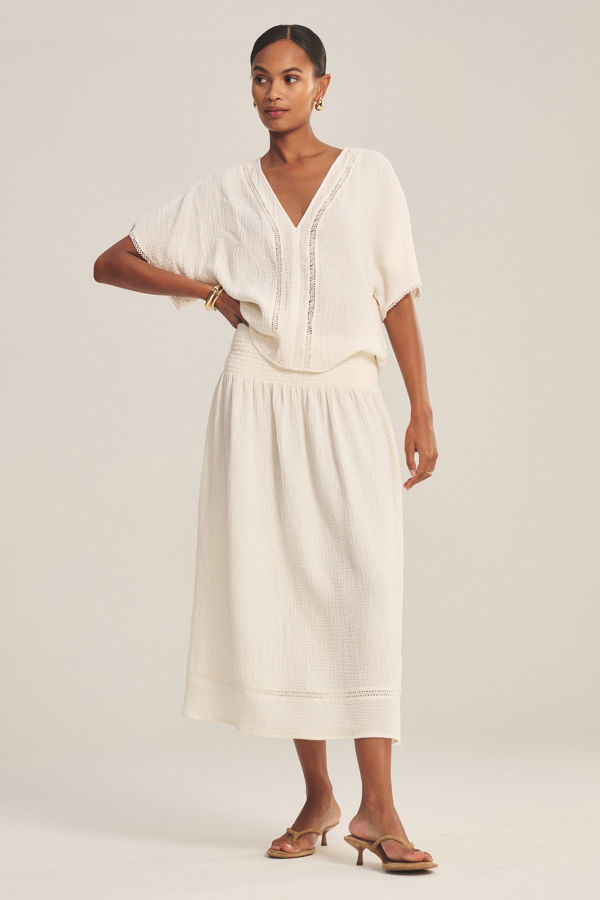 A woman wears the MAZIE SKIRT by Velvet by Graham & Spencer—a cream cotton gauze skirt with a smocked waist—paired with a matching short-sleeve blouse, sandals, pulled-back hair, gold earrings, and bracelets.-41296919986369