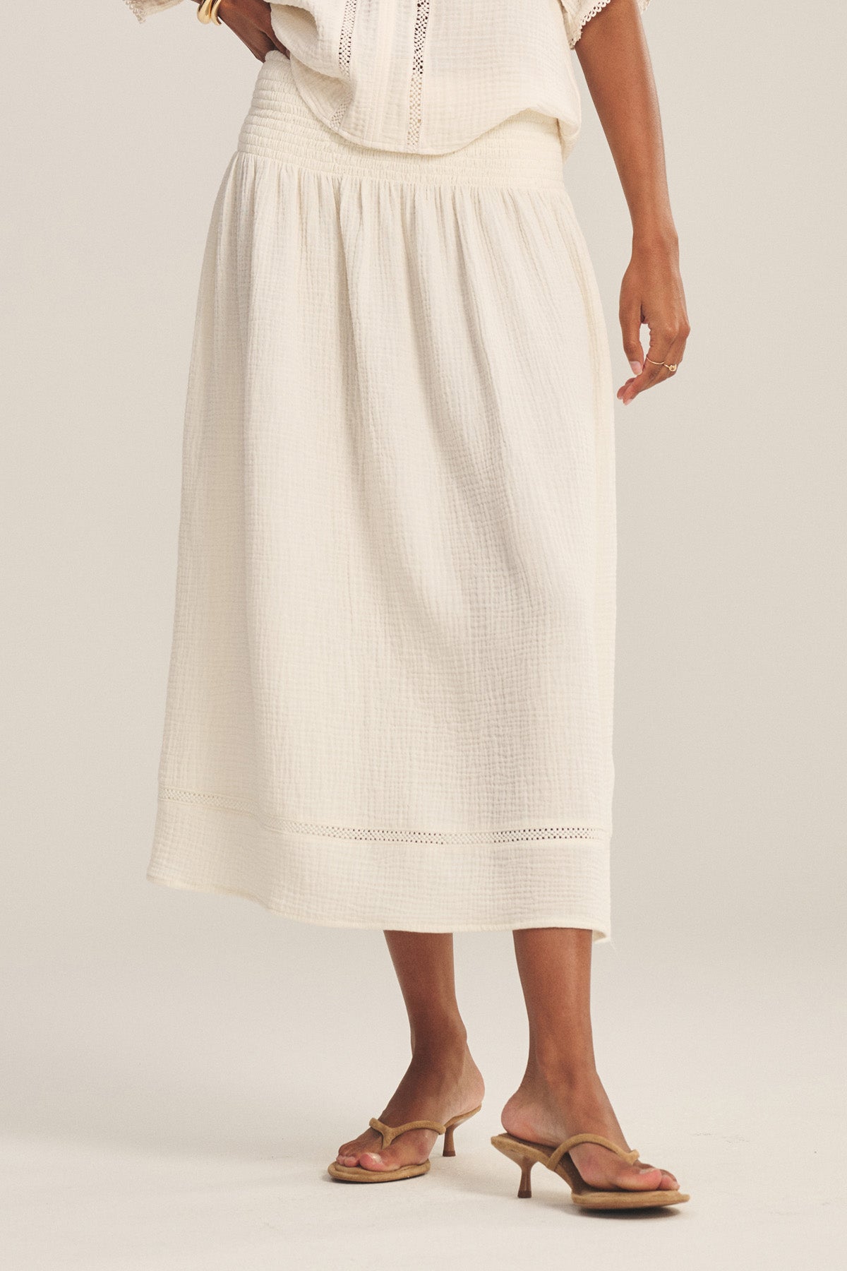 A person wearing the MAZIE SKIRT by Velvet by Graham & Spencer—a white cotton gauze skirt with a textured finish—paired with a white top and heeled sandals stands against a neutral background.