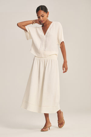 A woman poses in a relaxed stance wearing the MAZIE SKIRT from Velvet by Graham & Spencer—a loose-fitting white cotton gauze skirt paired with a matching white short-sleeve blouse and tan sandals against a plain background.