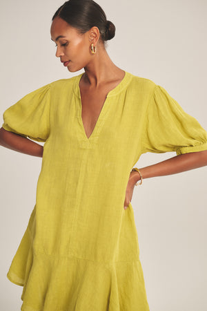 A woman in the MEADOW DRESS by Velvet by Graham & Spencer—a yellow woven linen dress with puff sleeves and a ruffle-tiered hem—paired with gold hoop earrings and a gold bracelet, stands against a plain background.