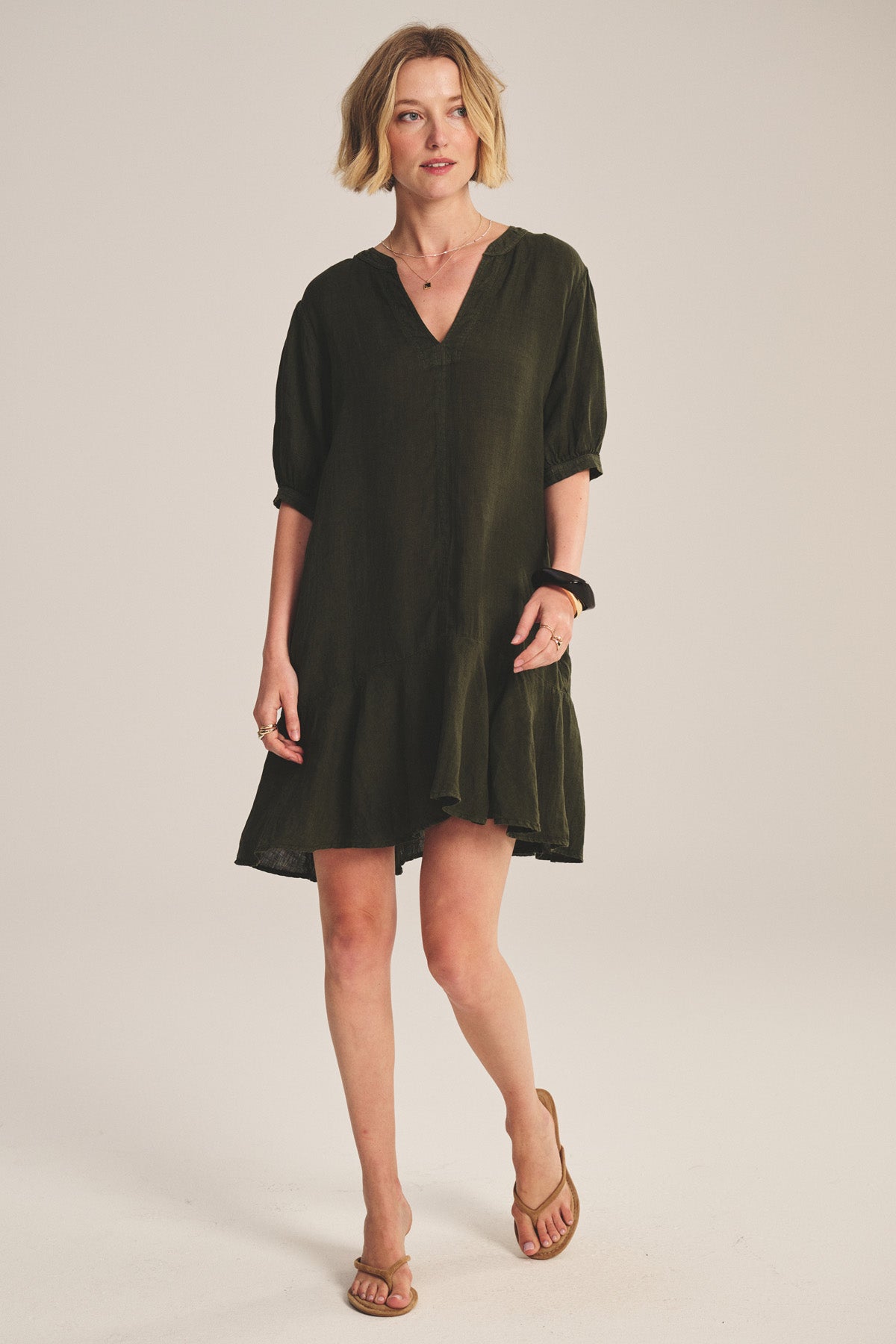 A woman wears the Velvet by Graham & Spencer MEADOW DRESS, a loose, knee-length woven linen style with a ruffle-tiered hem, paired with tan sandals against a plain background.
