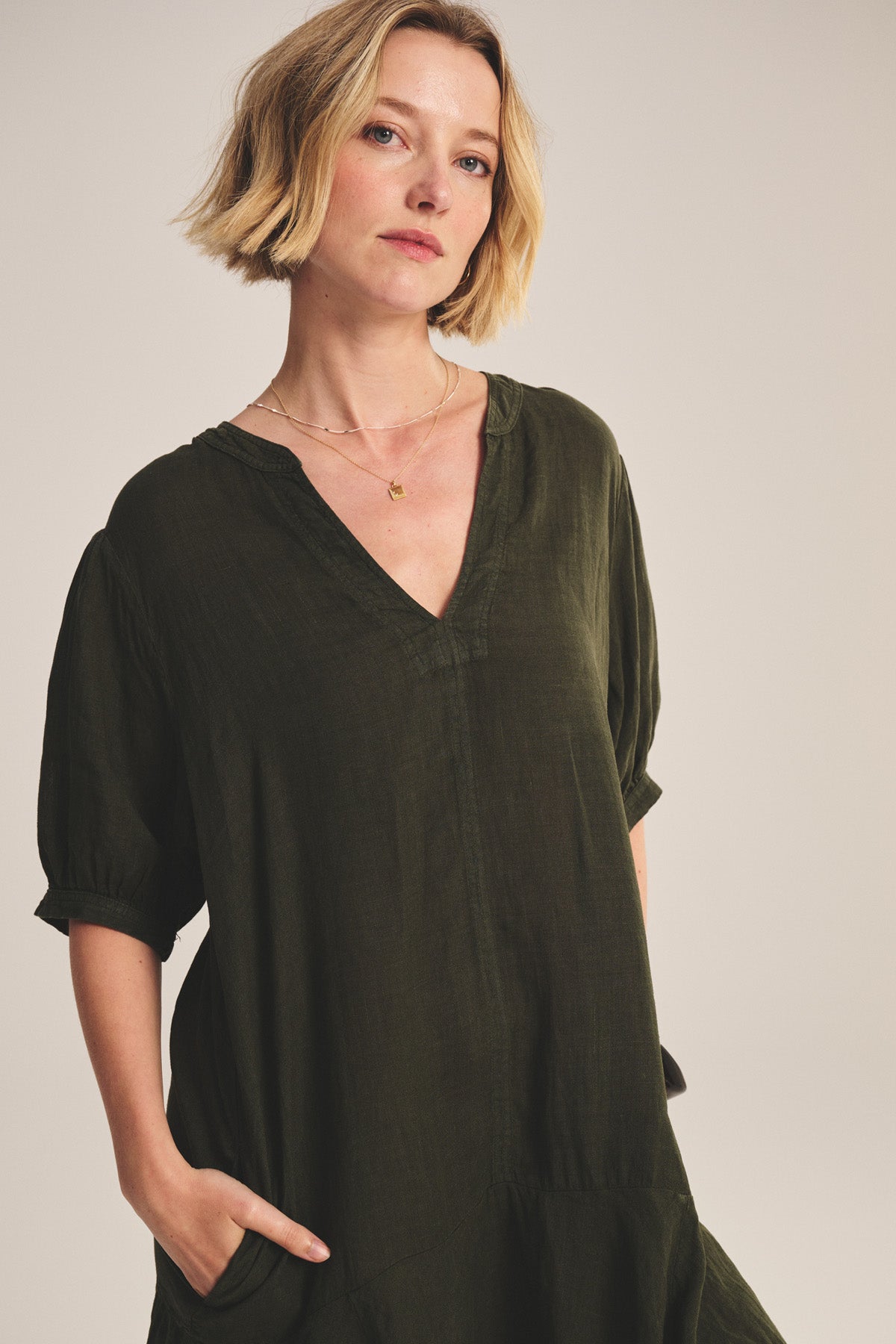 A woman wears the MEADOW DRESS by Velvet by Graham & Spencer, a loose dark green linen dress featuring short puff sleeves, a V-neck, and pockets, standing against a plain background.