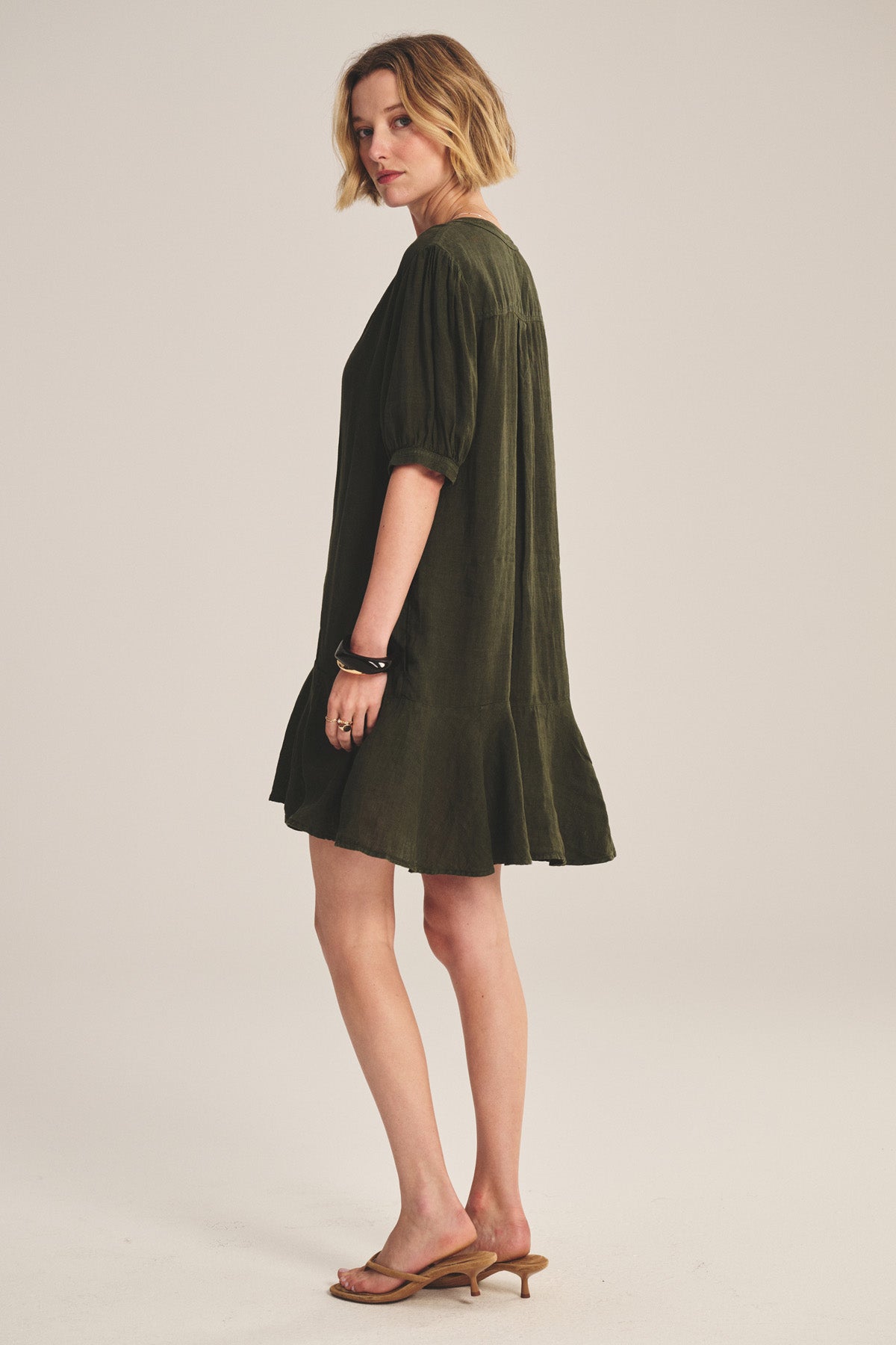 A woman stands sideways against a plain background wearing the MEADOW DRESS by Velvet by Graham & Spencer—a loose-fitting dark green linen dress with a ruffle-tiered hem—paired with tan heeled sandals.