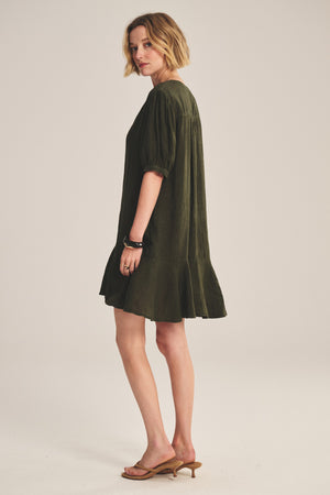 A woman stands sideways against a plain background wearing the MEADOW DRESS by Velvet by Graham & Spencer—a loose-fitting dark green linen dress with a ruffle-tiered hem—paired with tan heeled sandals.