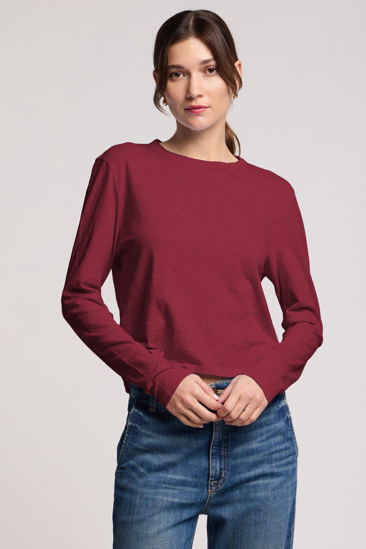 A person wears the MEGAN TEE by Velvet by Graham & Spencer, a maroon long-sleeve textured cotton slub top, paired with blue jeans, standing against a plain background.