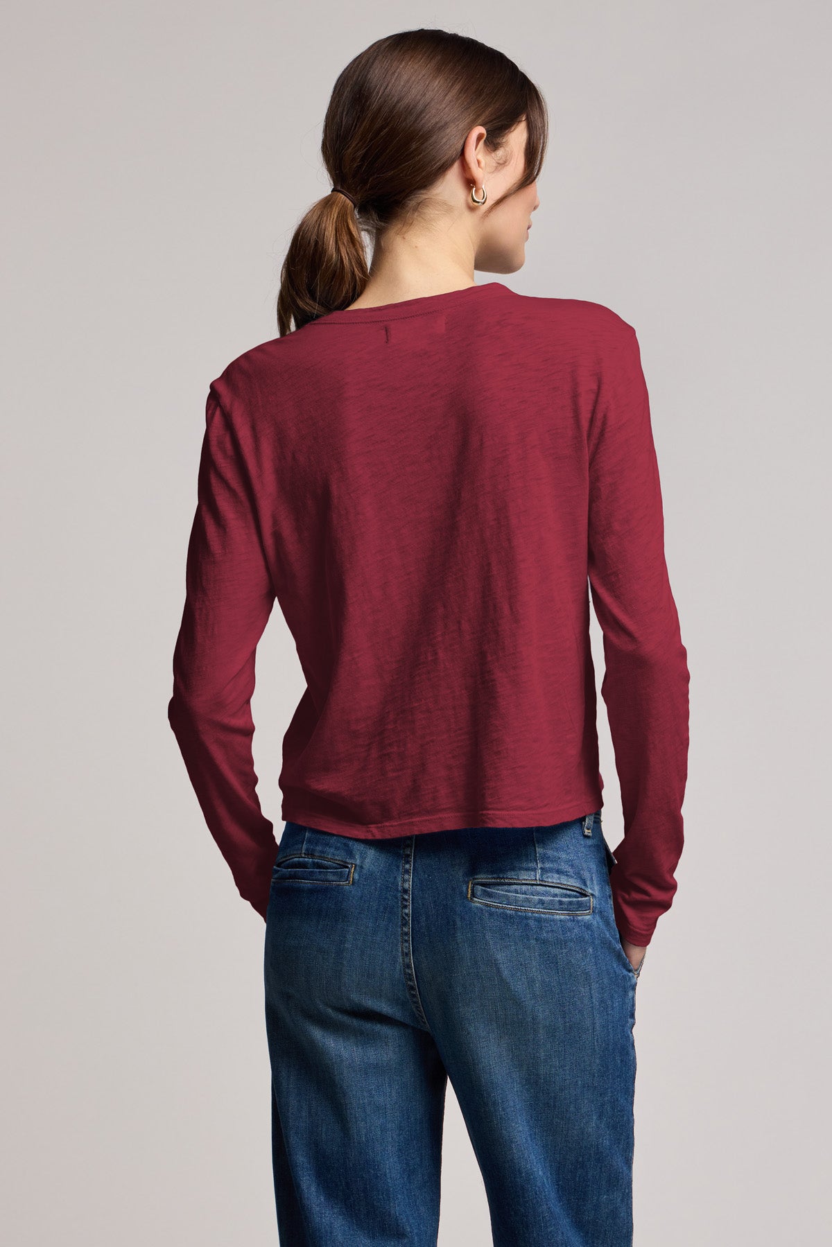 A woman with brown hair in a low ponytail models the MEGAN TEE by Velvet by Graham & Spencer in burgundy, paired with blue jeans. She shows off its cropped fit as she faces away from the camera against a plain background.-40430945435841