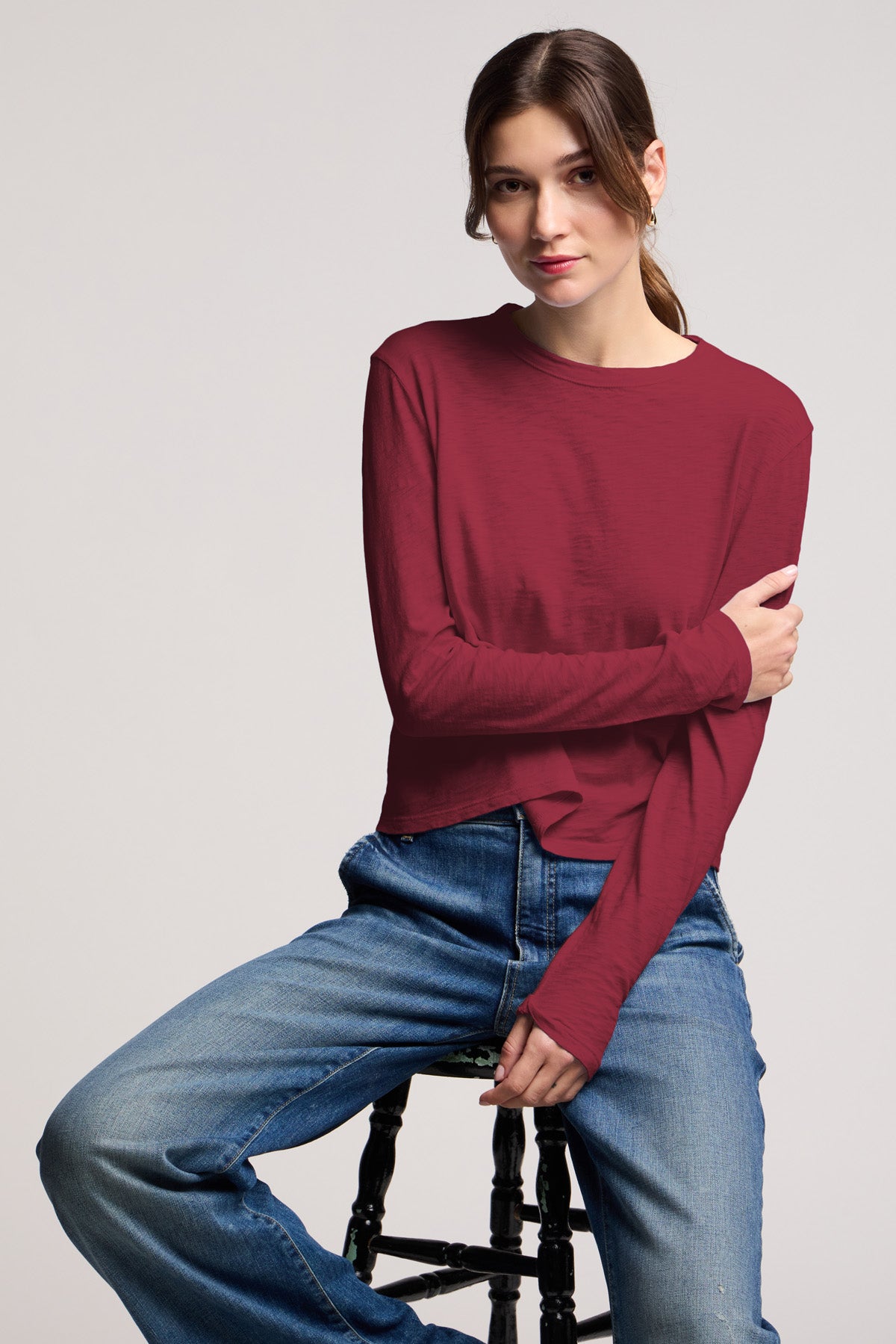 A woman sits on a stool against a plain background, wearing the Velvet by Graham & Spencer MEGAN TEE in maroon (cropped fit, long sleeves) with blue jeans, one arm crossed over her body.