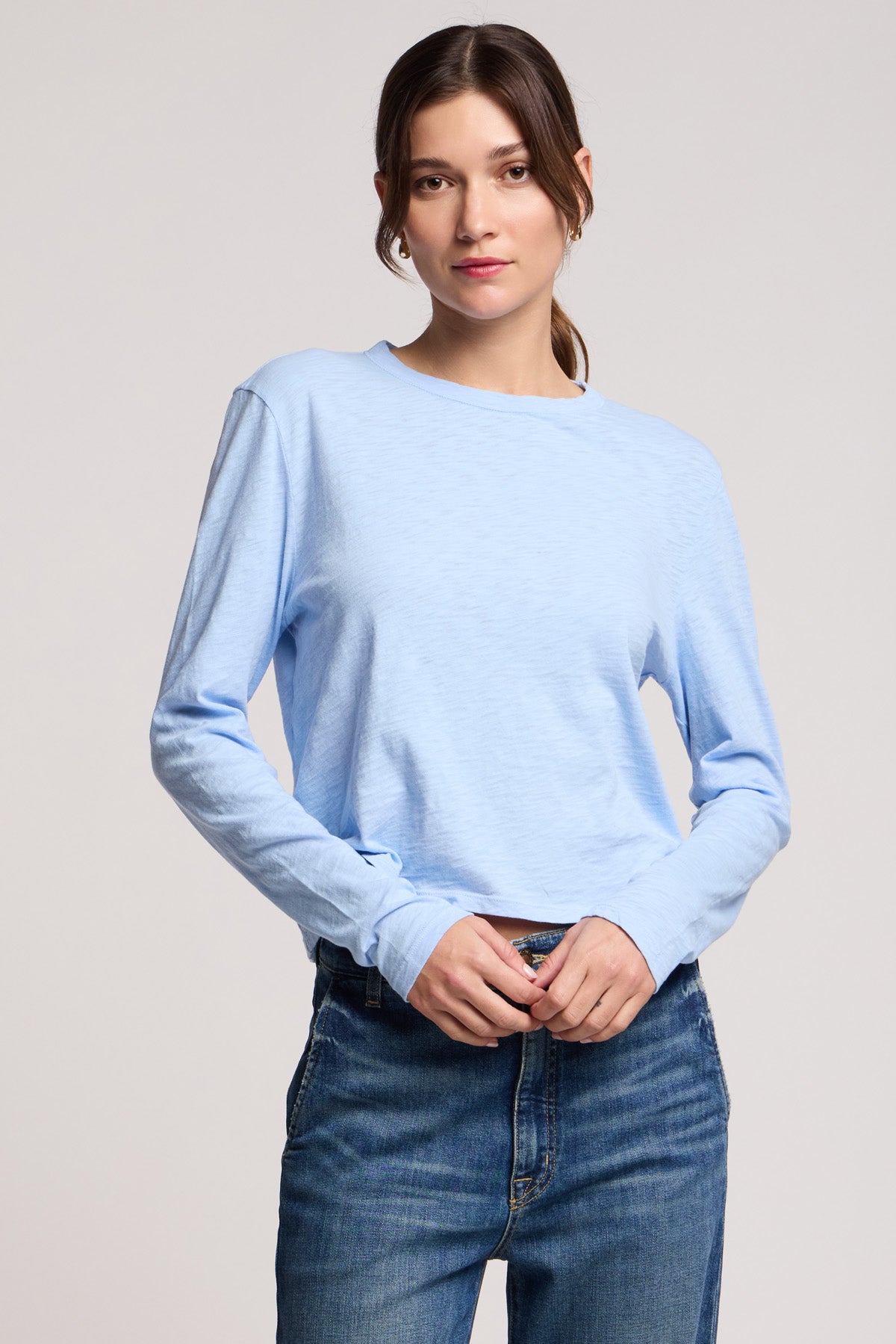 A woman faces forward against a plain background, wearing the MEGAN TEE by Velvet by Graham & Spencer—a light blue, long-sleeve, cropped-fit top—paired with blue jeans.-40329283076289