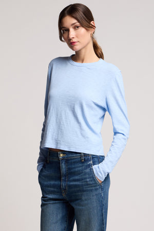 A woman wears the MEGAN TEE by Velvet by Graham & Spencer—a light blue, relaxed-fit, long-sleeve cropped tee—with blue jeans, standing with one hand in her pocket against a plain background.