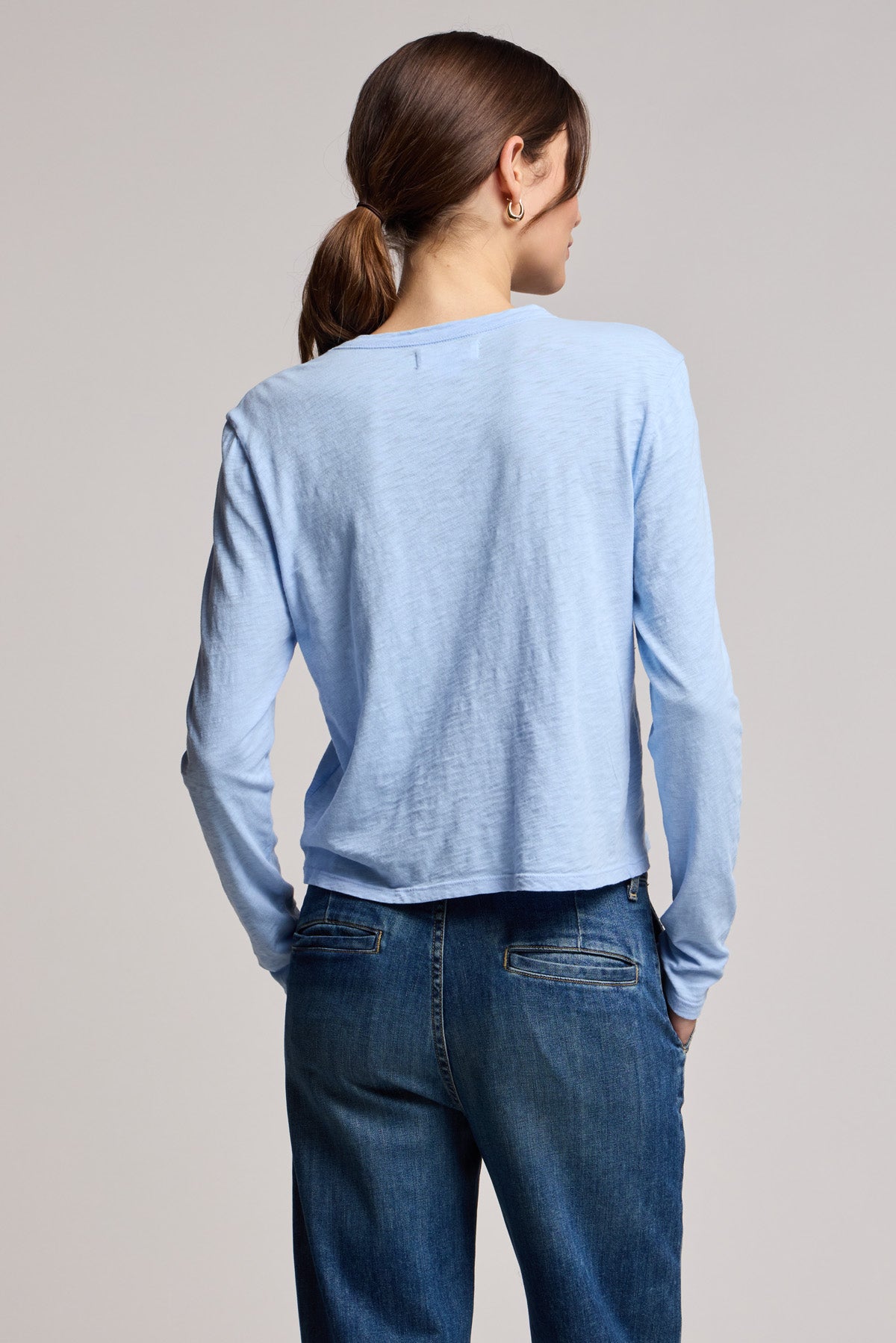 A person with brown hair in a low ponytail wears the MEGAN TEE by Velvet by Graham & Spencer—a light blue cotton slub long-sleeve—paired with blue jeans, standing with their back to the camera.-40329283010753