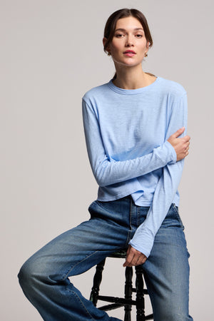 A person with light skin and brown hair wears the MEGAN TEE by Velvet by Graham & Spencer (light blue, classic fit, long sleeves) and blue jeans while seated on a black stool against a plain background.