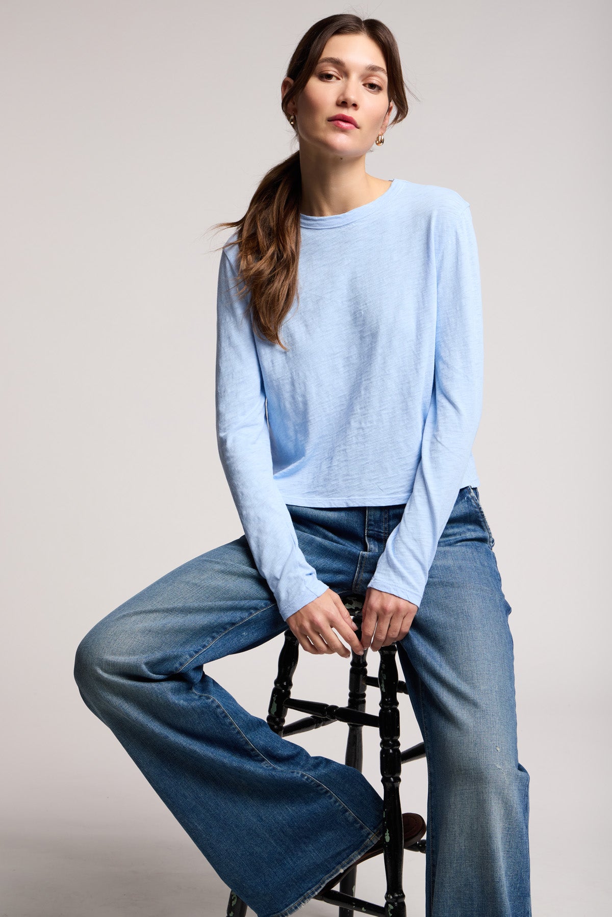 A person with long brown hair, wearing the MEGAN TEE by Velvet by Graham & Spencer and wide-leg jeans, sits on a black stool against a plain background.-40329282846913