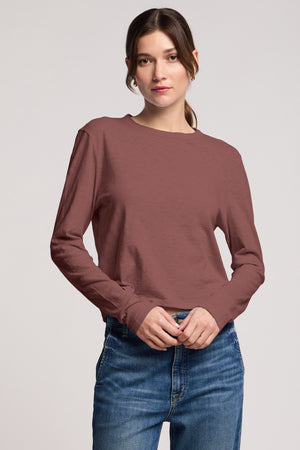 A woman faces forward, wearing the MEGAN TEE by Velvet by Graham & Spencer—a cropped fit long-sleeve brown top—paired with blue jeans against a plain light background.