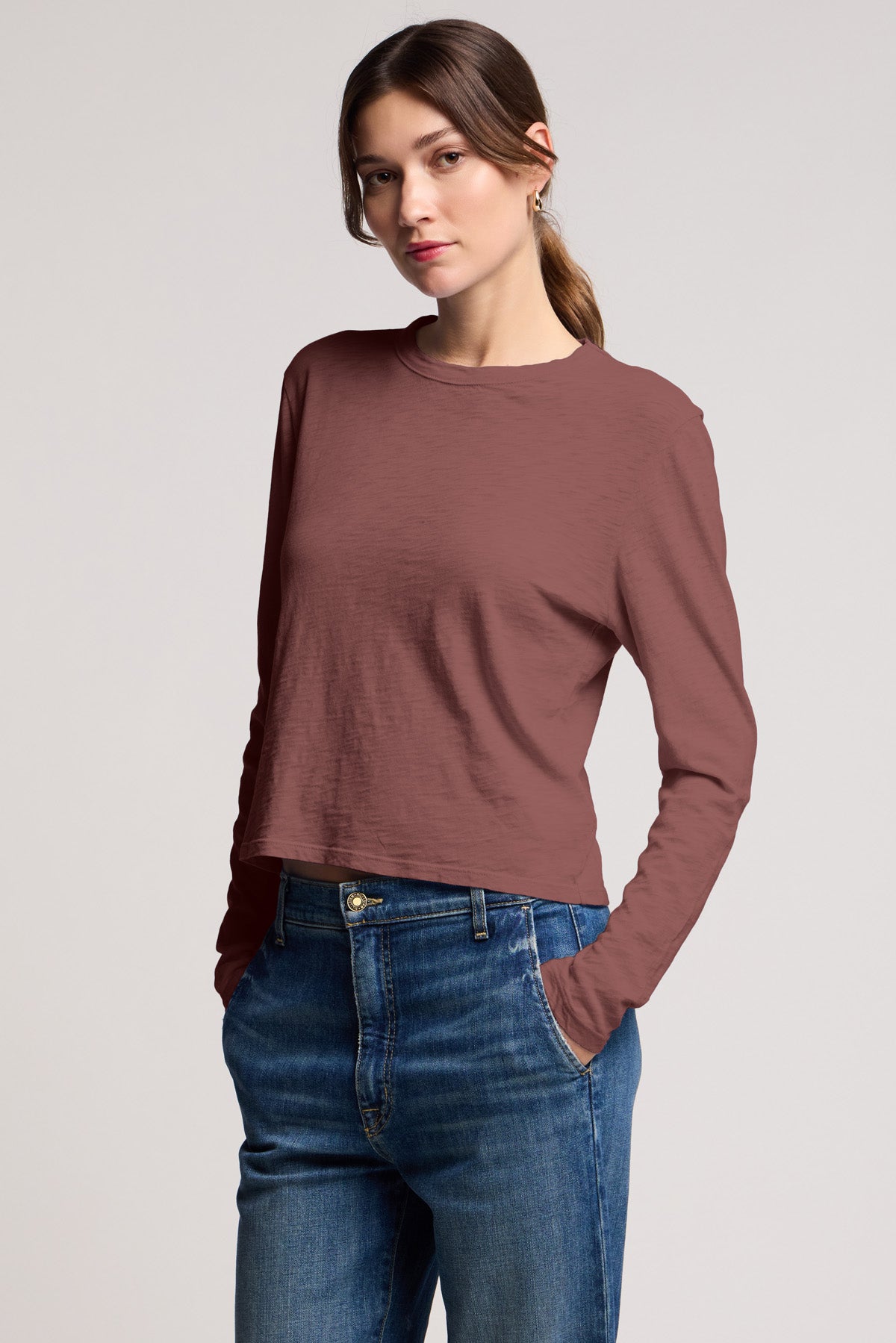 A woman stands with hands in pockets, wearing the Velvet by Graham & Spencer MEGAN TEE—a mauve long-sleeve crop top—paired with blue jeans, set against a plain light background.