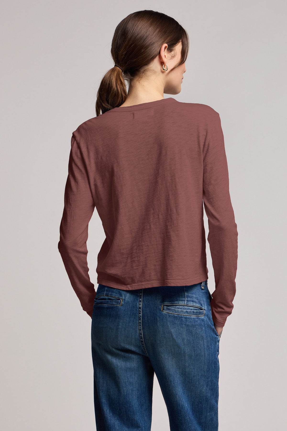 A person with brown hair in a low ponytail stands with their back to the camera, wearing the MEGAN TEE by Velvet by Graham & Spencer—a cropped, long-sleeve top—paired with blue jeans.-40430950580417