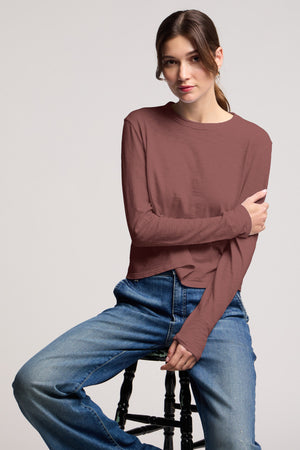 A woman faces the camera, hair tied back and one arm crossed, wearing the MEGAN TEE by Velvet by Graham & Spencer—a cropped long-sleeve top—paired with blue jeans while sitting on a stool.