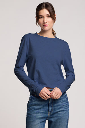 A woman stands against a plain background wearing the MEGAN TEE in blue textured cotton slub and blue jeans from Velvet by Graham & Spencer, looking directly at the camera with a neutral expression.