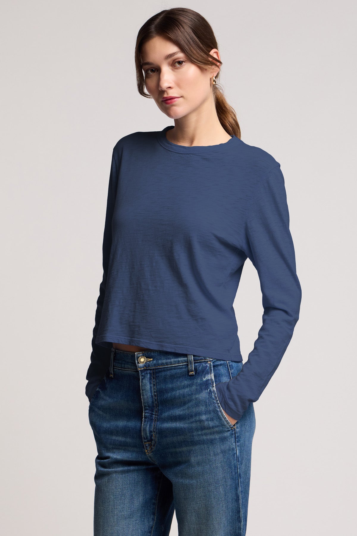 A woman stands with one hand in her pocket, wearing the cropped MEGAN TEE by Velvet by Graham & Spencer and blue jeans against a plain light background.
