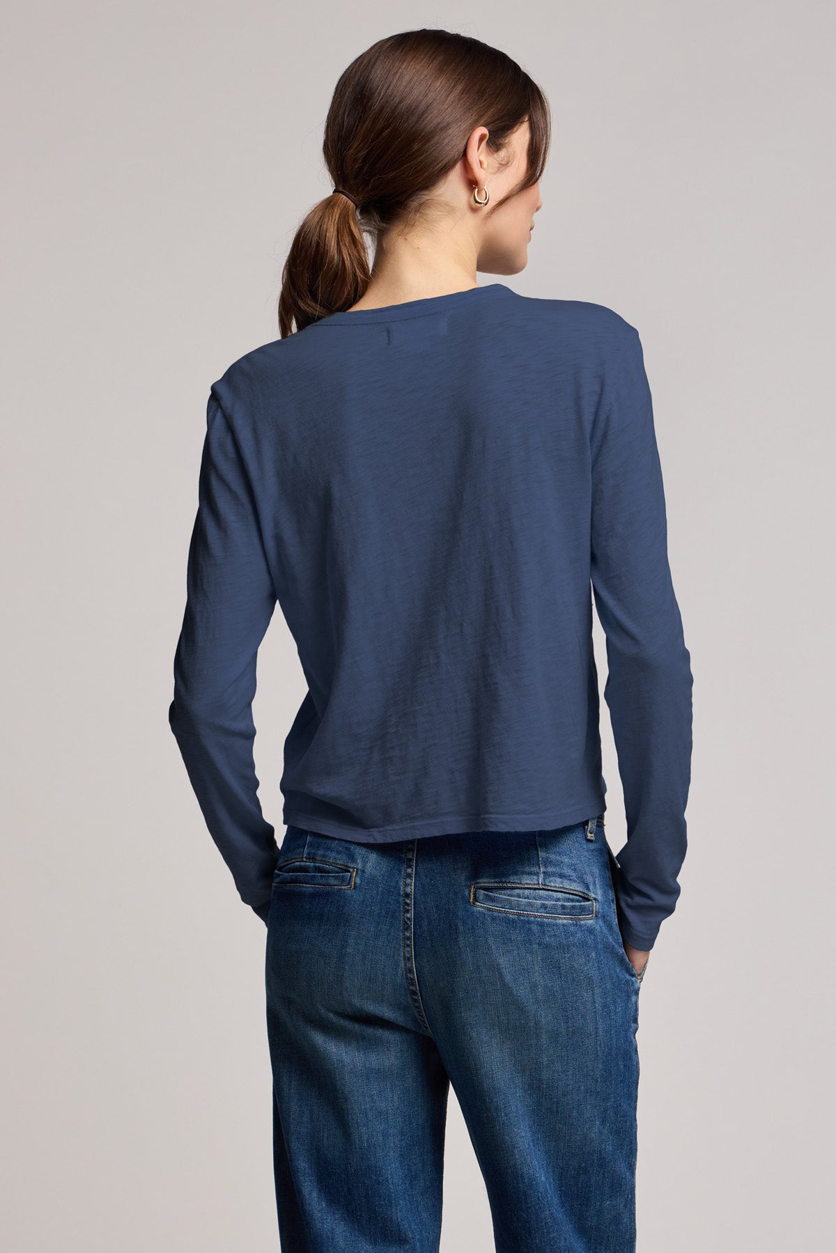 A person with brown hair in a low ponytail, wearing the MEGAN TEE by Velvet by Graham & Spencer and blue jeans, stands with their back to the camera.