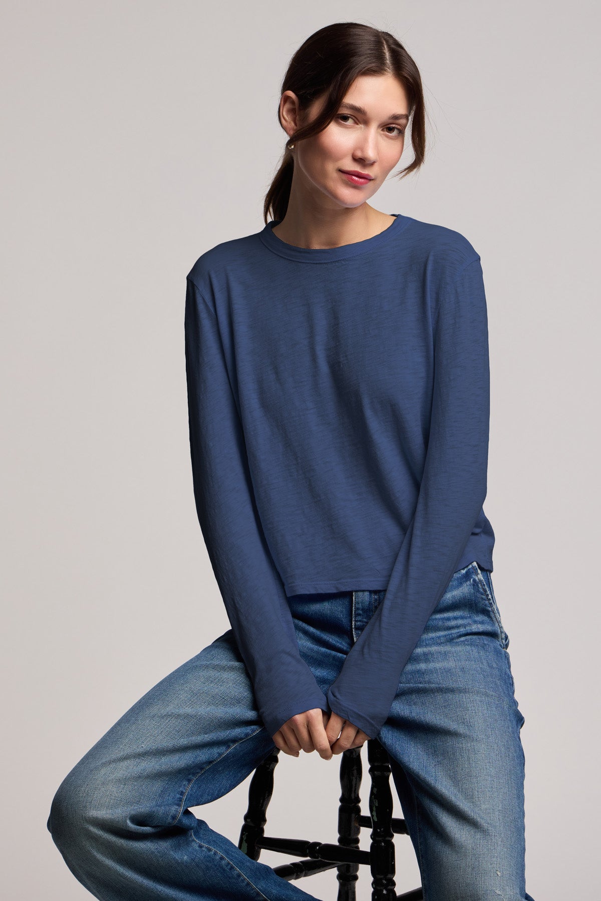 A person wearing the MEGAN TEE by Velvet by Graham & Spencer, a blue long-sleeve textured cotton slub top, paired with jeans, sits on a stool against a plain background.-40430950777025