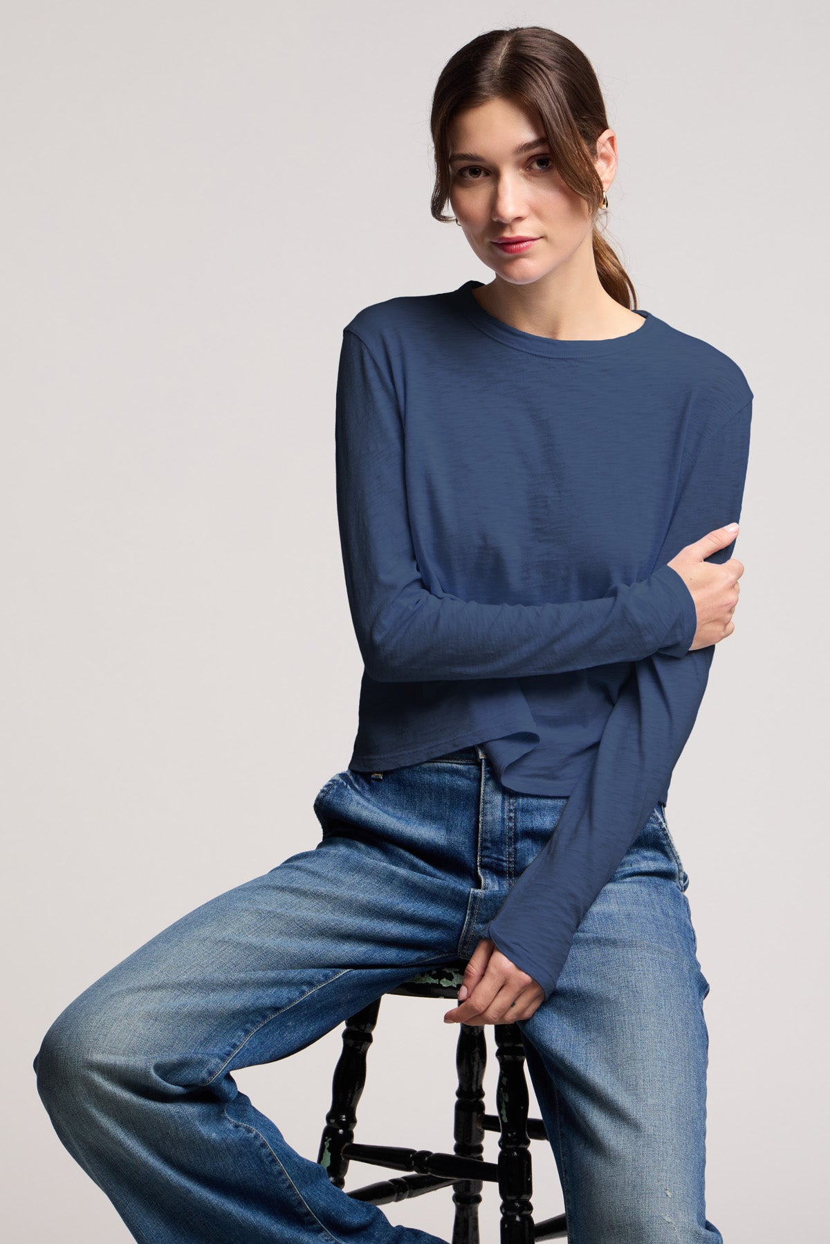 A woman with dark hair in a low ponytail sits on a stool against a plain light background, wearing the MEGAN TEE by Velvet by Graham & Spencer and blue jeans.-40430950809793