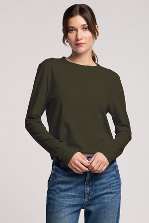 A woman stands against a plain background wearing the MEGAN TEE by Velvet by Graham & Spencer in olive green textured cotton slub, paired with blue jeans and her hands clasped in front.