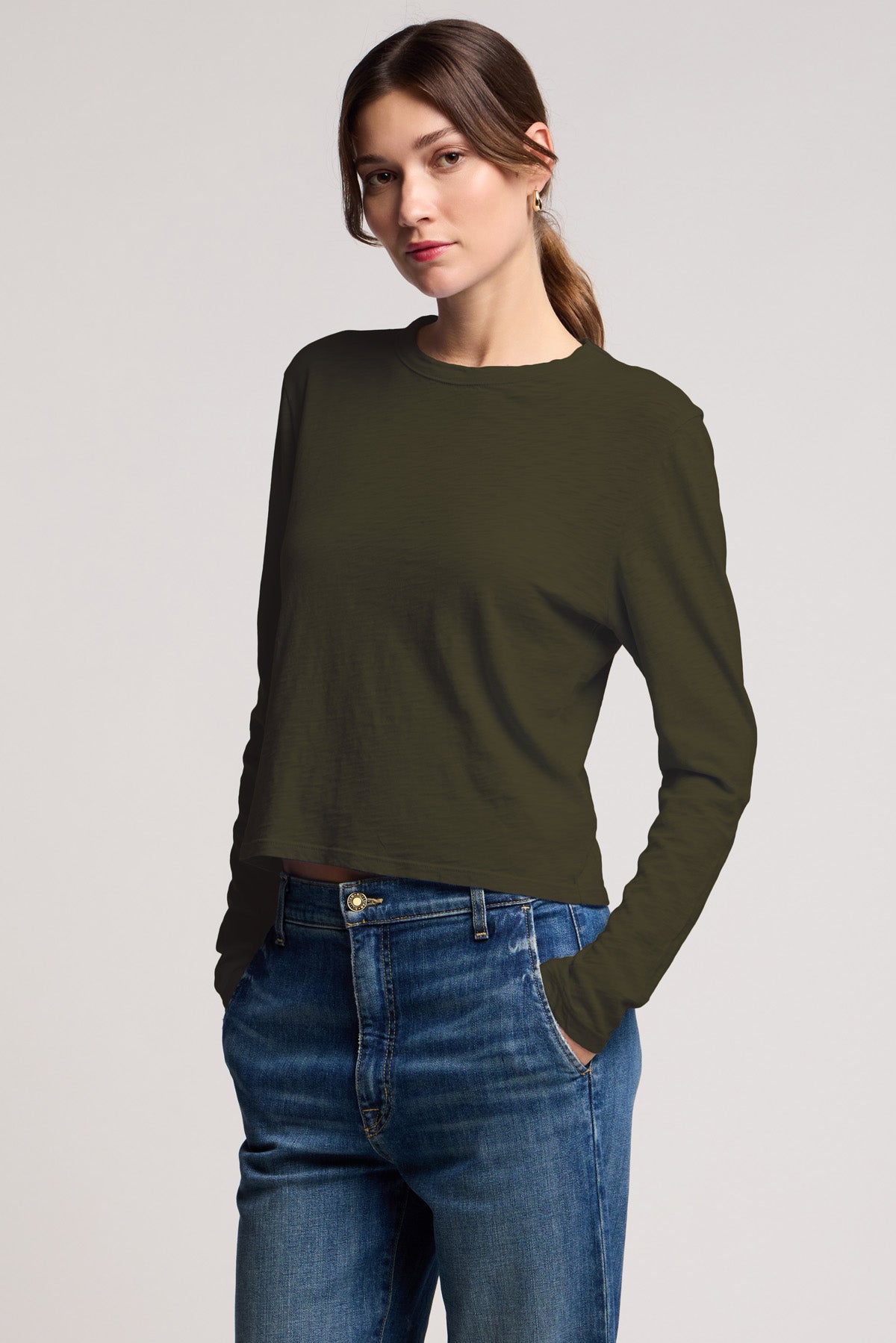 A woman stands with her hands in her jeans pockets, wearing the MEGAN TEE by Velvet by Graham & Spencer—a long-sleeve, dark green textured cotton slub tee—against a plain light background.-40808429322433