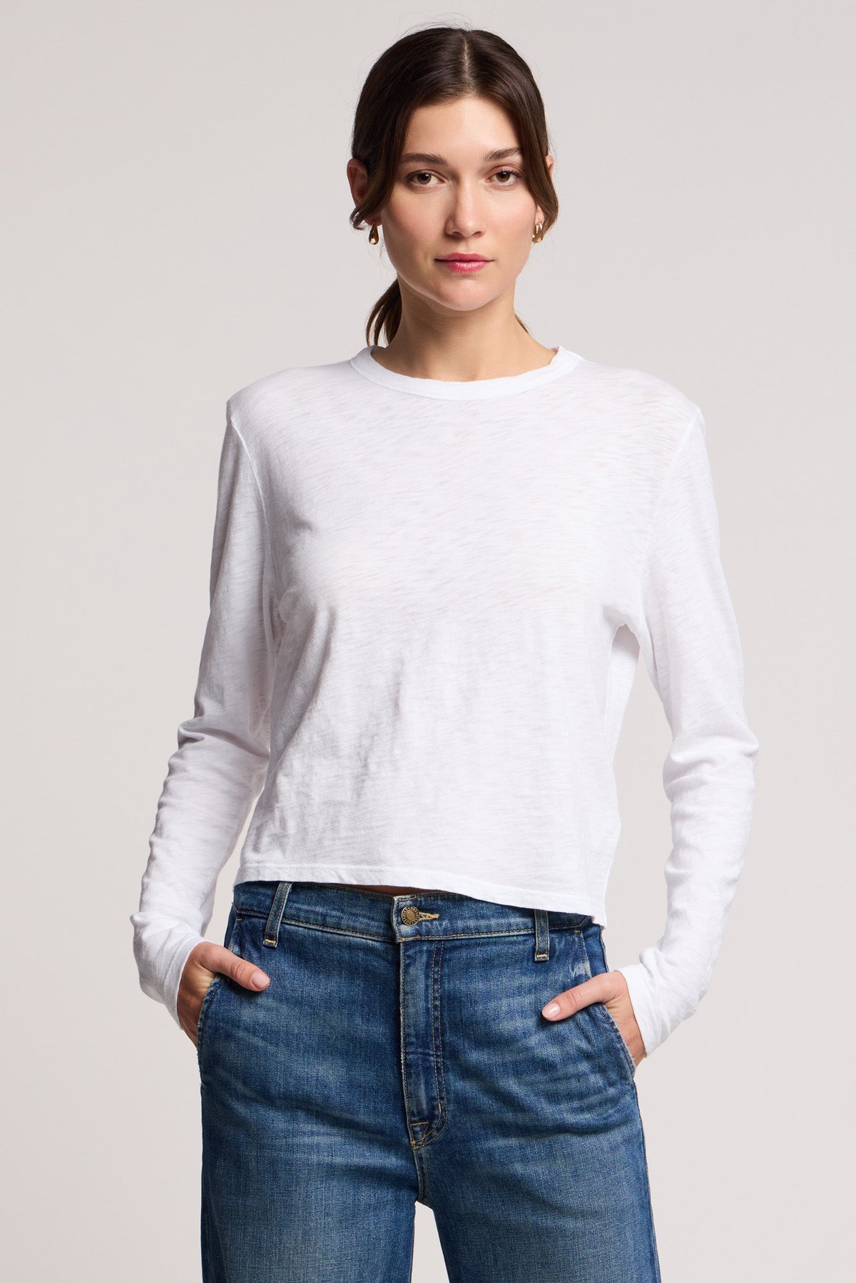 A woman stands facing forward with hands in pockets, wearing the MEGAN TEE by Velvet by Graham & Spencer, a white cotton slub long-sleeve top, paired with blue jeans against a plain light background.-40329282781377