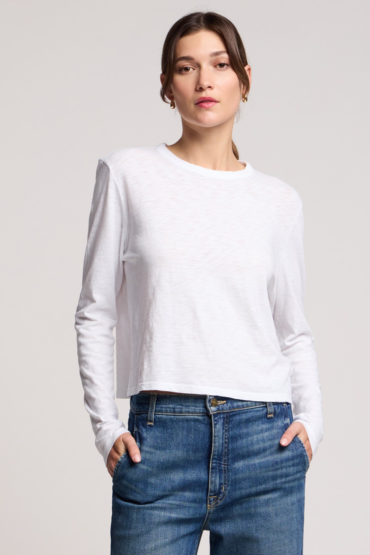 A person wearing the MEGAN TEE by Velvet by Graham & Spencer, a plain white cotton slub long-sleeve shirt, and blue jeans stands with hands in pockets against a neutral background.-40329282748609