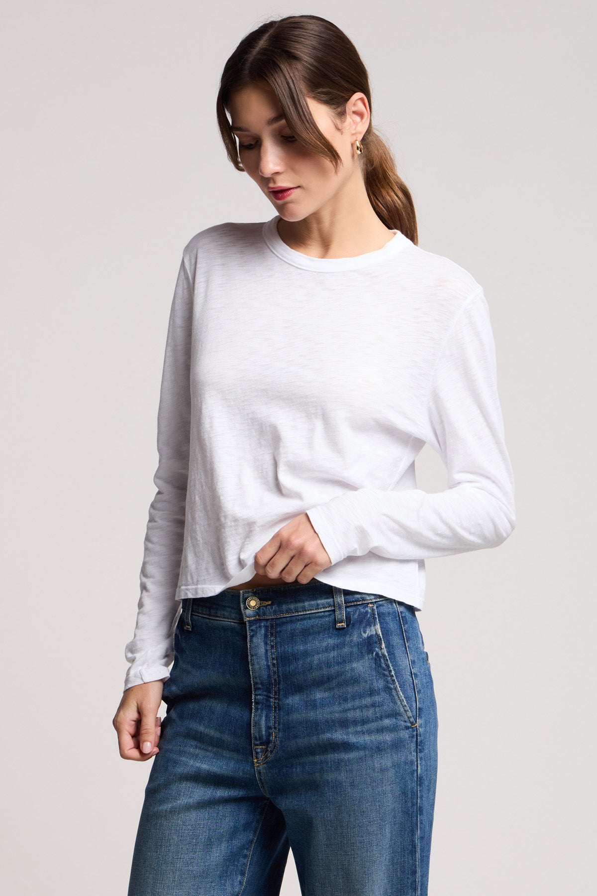 A woman wearing the MEGAN TEE by Velvet by Graham & Spencer with blue jeans stands against a neutral background, looking downward.-40329282715841