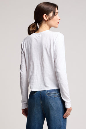 A woman with brown hair in a low ponytail stands with her back to the camera against a plain background, wearing the MEGAN TEE by Velvet by Graham & Spencer—a cropped fit long-sleeve tee—paired with blue jeans.