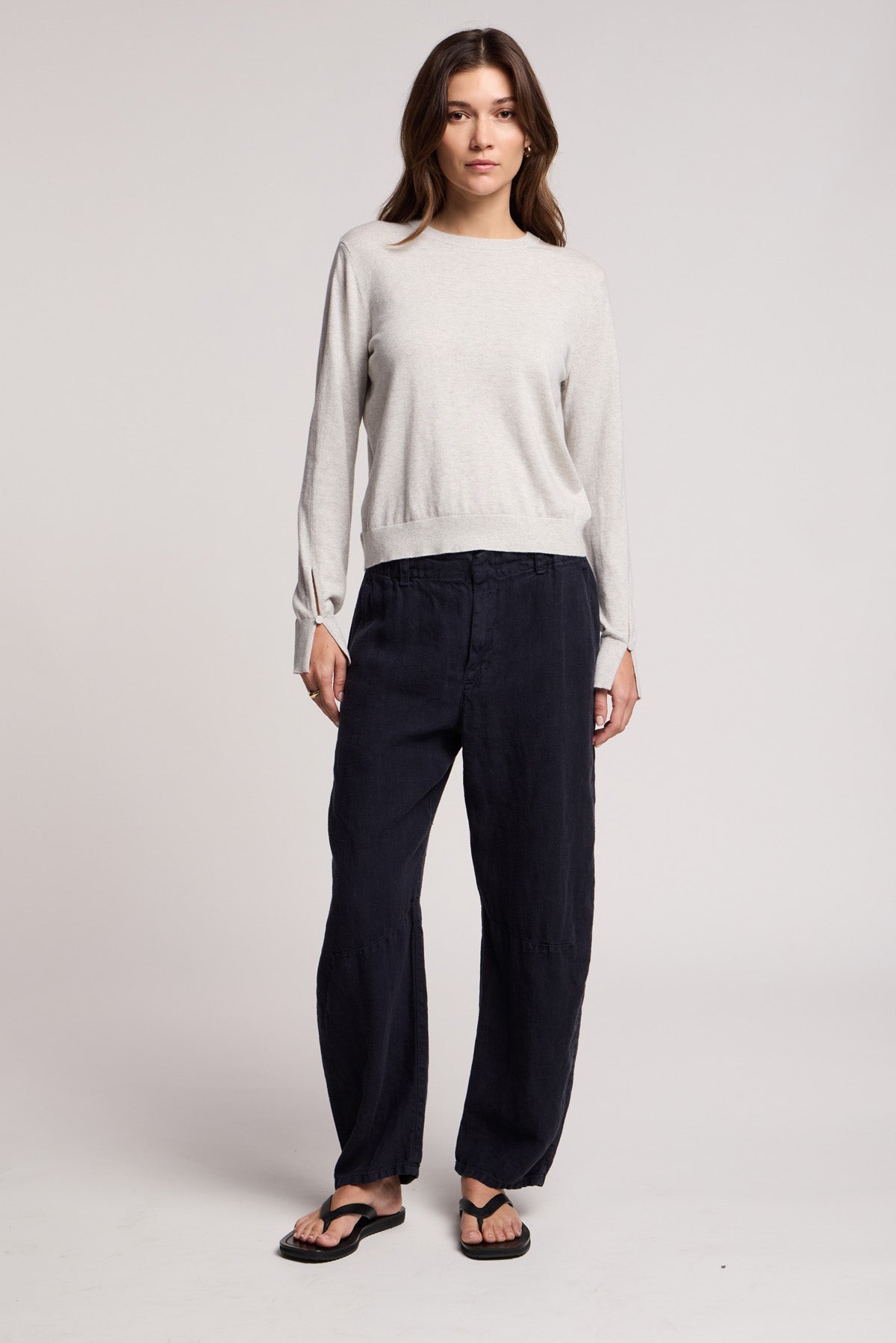A woman faces forward wearing the MELINA SWEATER by Velvet by Graham & Spencer—a light gray crew neck with ribbed trims—paired with loose dark pants and black sandals, standing against a plain background.-40672322846913