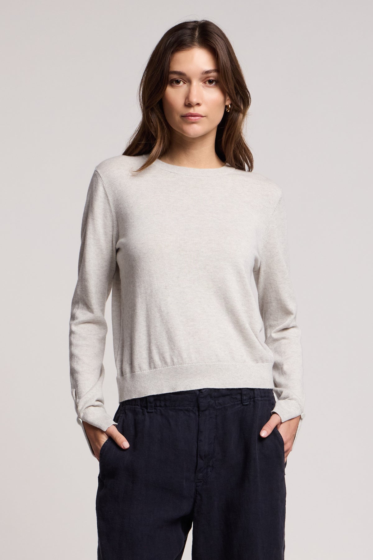 A woman stands facing forward with hands in pockets, wearing the MELINA SWEATER by Velvet by Graham & Spencer—a light grey cotton-cashmere piece with ribbed trims—and dark pants against a plain background.