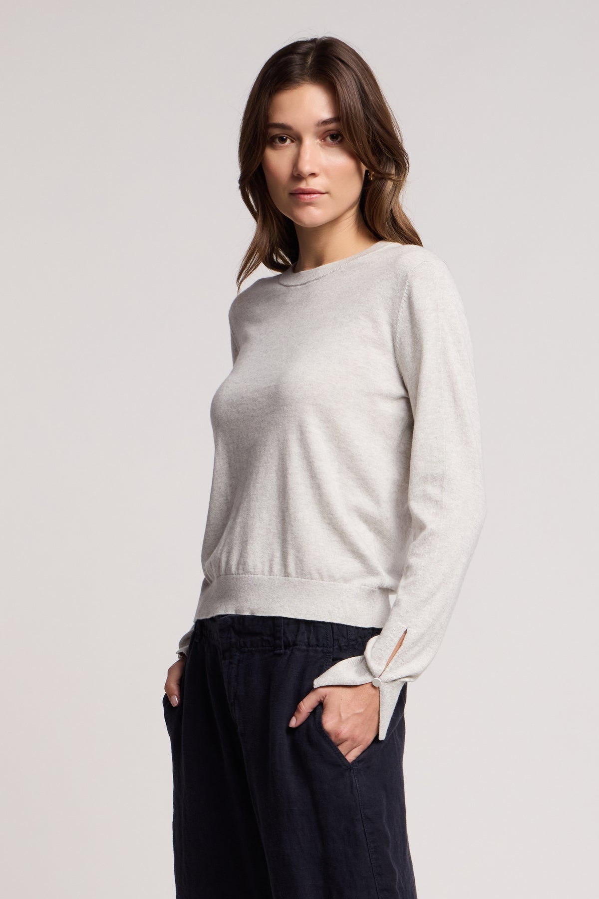 A woman faces forward with her left hand in her pocket, wearing the MELINA SWEATER by Velvet by Graham & Spencer—a light gray crew neck with ribbed trims—and dark pants against a plain background.