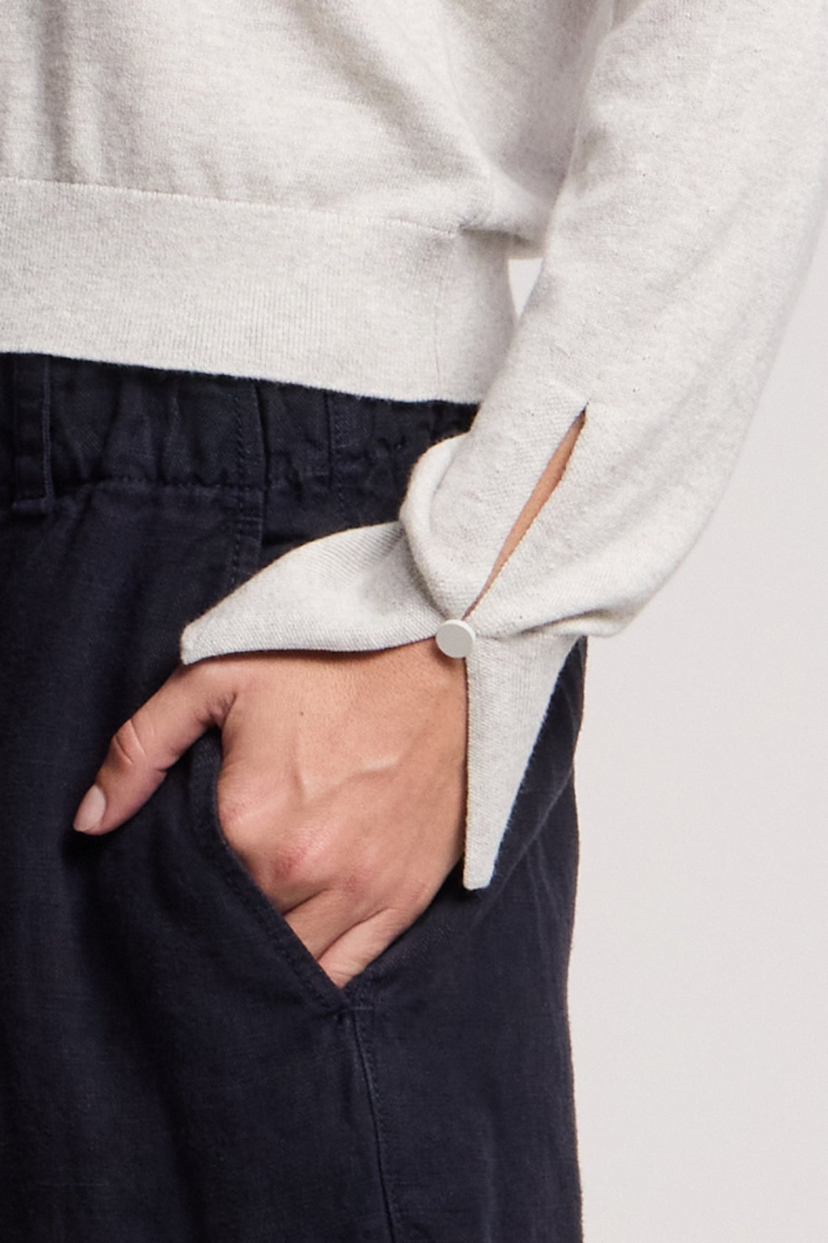 A person with a hand in their pocket wears the MELINA SWEATER by Velvet by Graham & Spencer—a light gray crew neck with ribbed, buttoned cuffs—paired with dark pants.-40672410861761