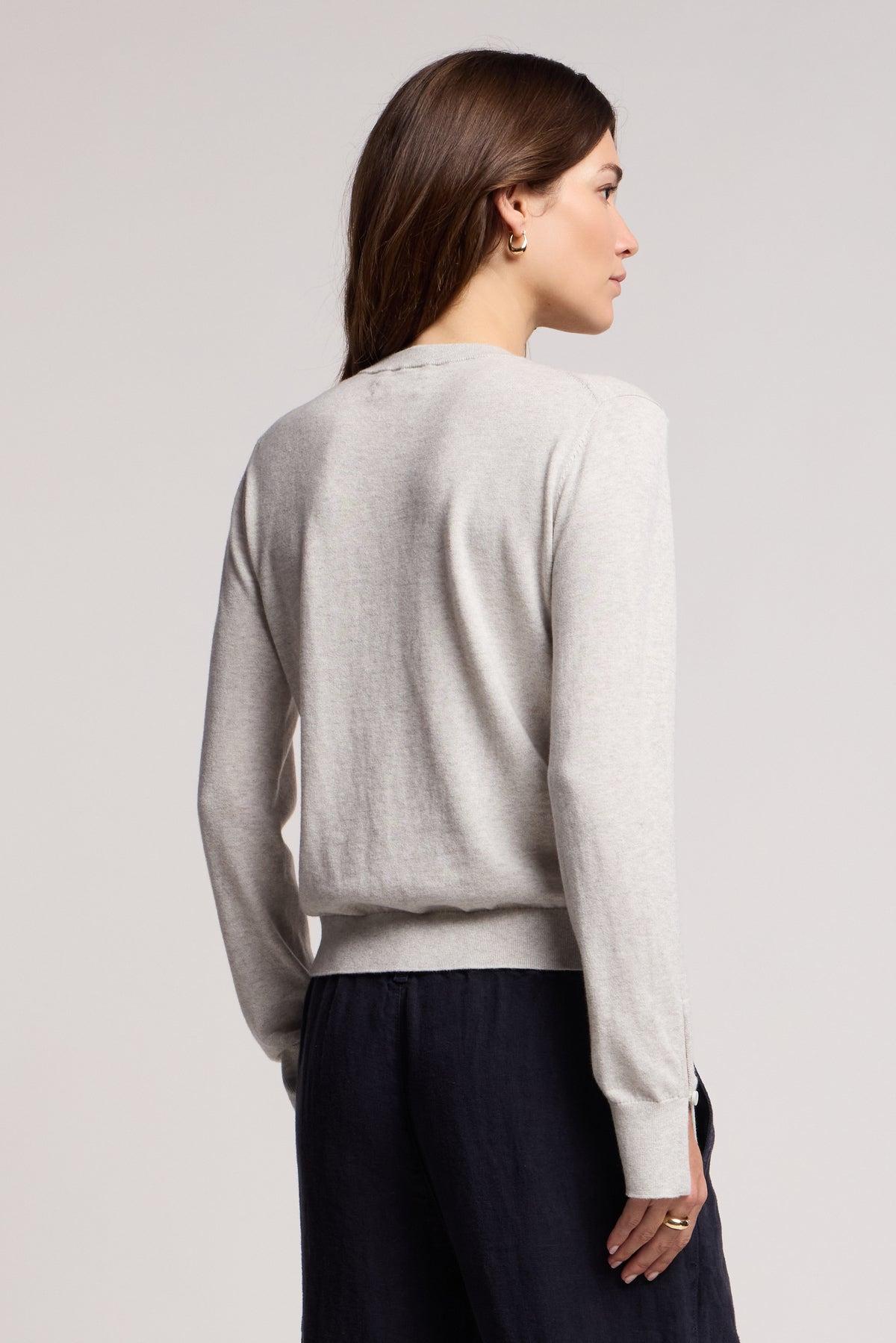 A woman stands with her back to the camera, wearing the MELINA SWEATER by Velvet by Graham & Spencer—a light gray cotton-cashmere crew neck with ribbed trims—paired with dark pants against a plain background.-40672410894529