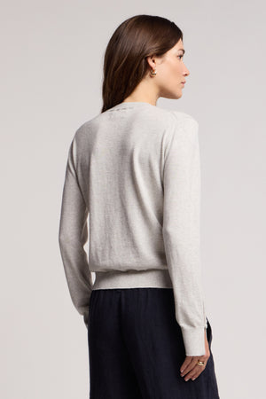 A woman stands with her back to the camera, wearing the MELINA SWEATER by Velvet by Graham & Spencer—a light gray cotton-cashmere crew neck with ribbed trims—paired with dark pants against a plain background.