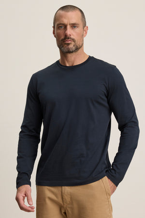 A man with short hair and a beard wears the ALFIE TEE by Velvet by Graham & Spencer in navy with beige pants, standing against a plain light background.