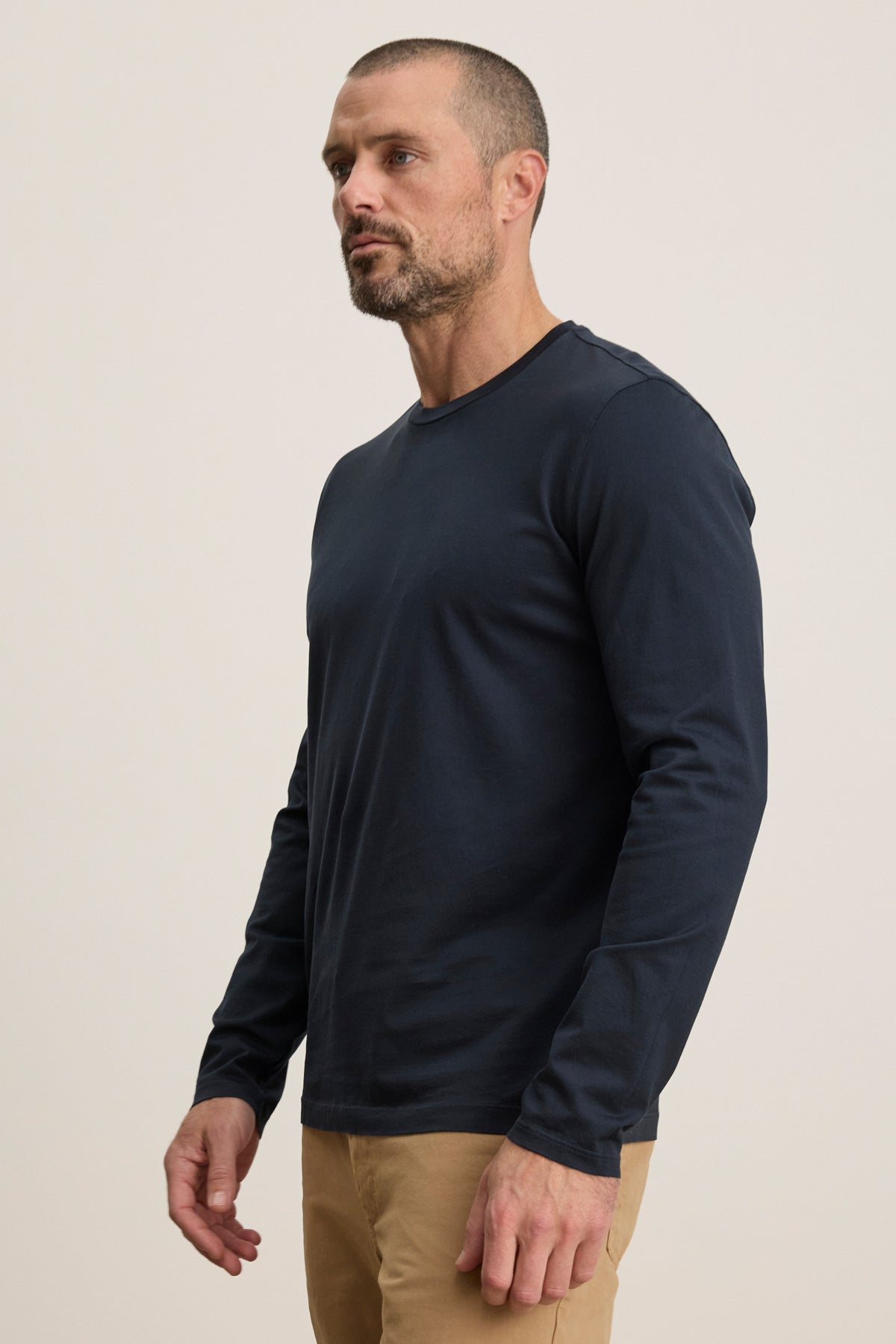   A man wears the ALFIE TEE by Velvet by Graham & Spencer, a navy blue midweight mercerized cotton long sleeve, with tan pants, standing against a plain light background, facing slightly to the side. 