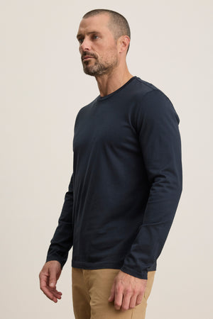 A man wears the ALFIE TEE by Velvet by Graham & Spencer, a navy blue midweight mercerized cotton long sleeve, with tan pants, standing against a plain light background, facing slightly to the side.