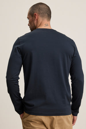 A man is seen from the back wearing the ALFIE TEE, a navy long sleeve tailored fit by Velvet by Graham & Spencer, with khaki pants and a bird tattoo visible on his neck.