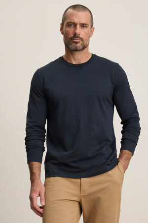A man with short hair and a beard wears the Velvet by Graham & Spencer ALFIE TEE, a navy tailored long sleeve shirt, paired with tan pants, standing against a plain light background.
