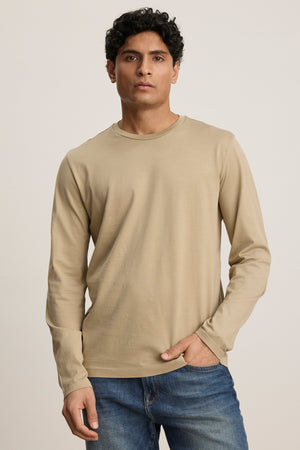 A man wearing the ALFIE TEE by Velvet by Graham & Spencer, a tailored fit beige long sleeve tee, and blue jeans stands against a plain light background.