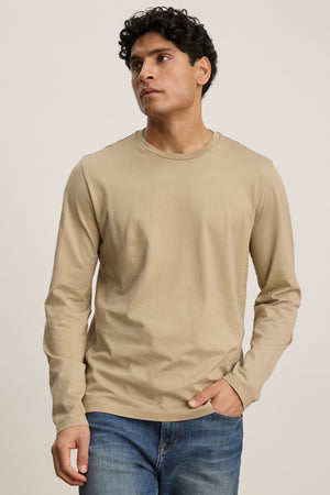 A man wearing the ALFIE TEE by Velvet by Graham & Spencer in beige with blue jeans stands against a plain light background, looking slightly to the side.