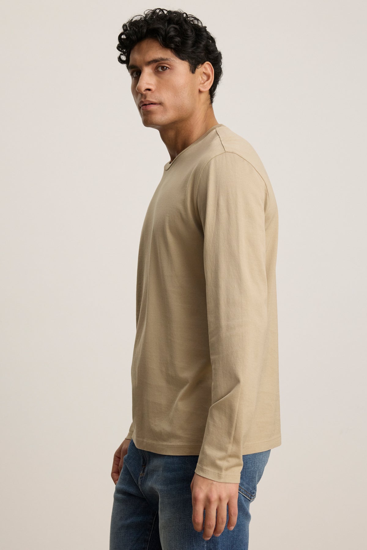 A man with short curly hair wears the ALFIE TEE by Velvet by Graham & Spencer in beige, styled with blue jeans, standing sideways against a plain light background.-39450512883905