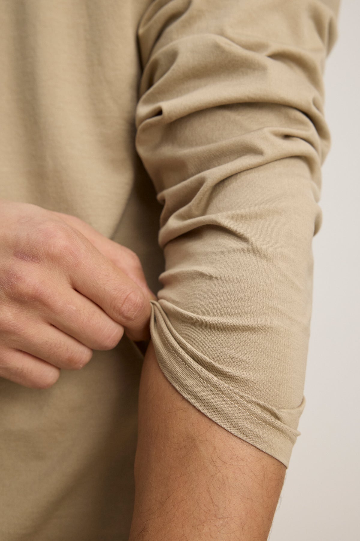 A person pulls up the sleeve of a beige Velvet by Graham & Spencer ALFIE TEE tailored fit long sleeve with their left hand.-39450512949441