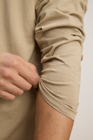 A person pulls up the sleeve of a beige Velvet by Graham & Spencer ALFIE TEE tailored fit long sleeve with their left hand.